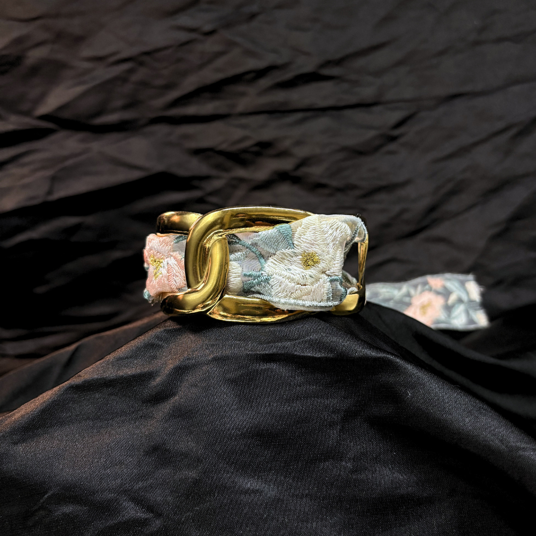 Gold thick link bracelet with a light blye and white lace woven through it on a black fabric background