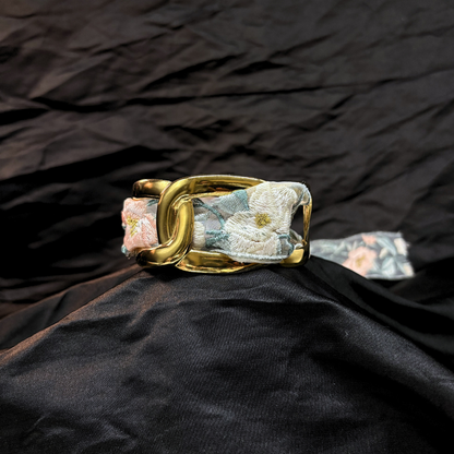 Gold thick link bracelet with a light blye and white lace woven through it on a black fabric background