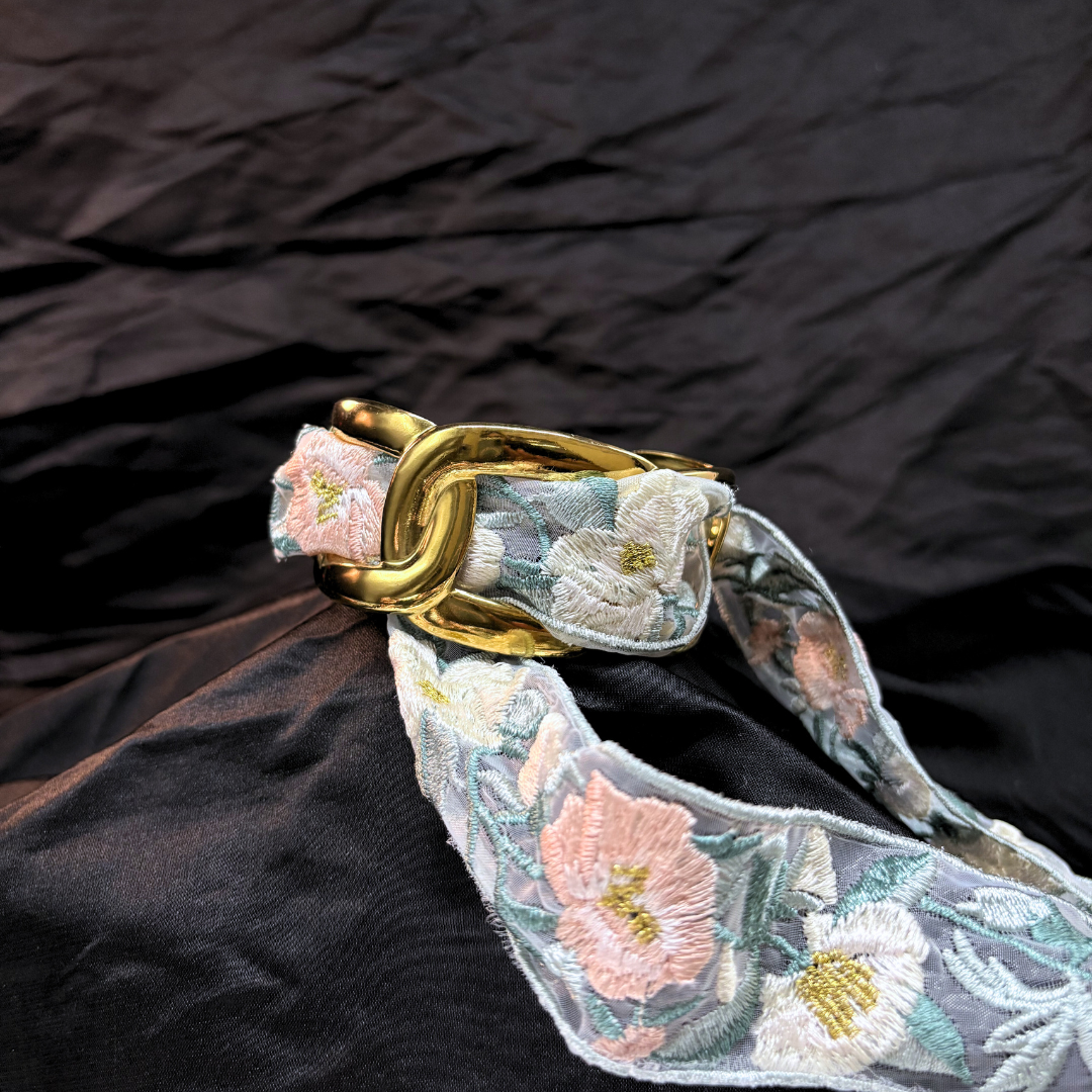 Gold thick link bracelet with a light blye and white lace woven through it on a black fabric background