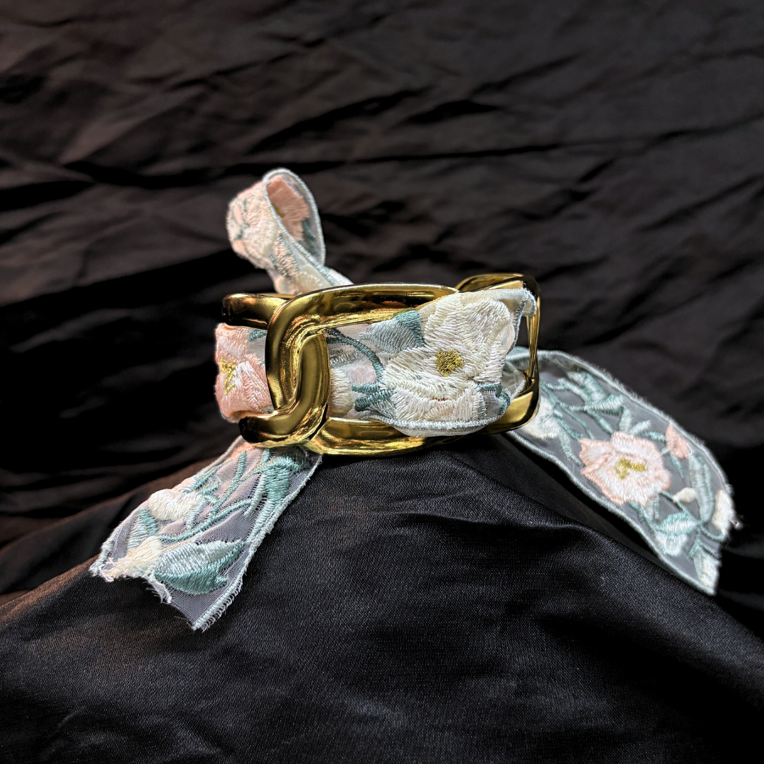 Gold thick link bracelet with a light blye and white lace woven through it on a black fabric background