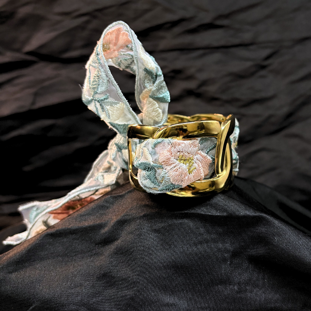 Gold thick link bracelet with a light blye and white lace woven through it on a black fabric background